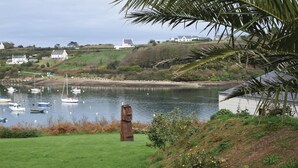 Une vue côtière avec des bateaux sur une étendue d’eau calme, un premier plan verdoyant et des maisons en arrière-plan.