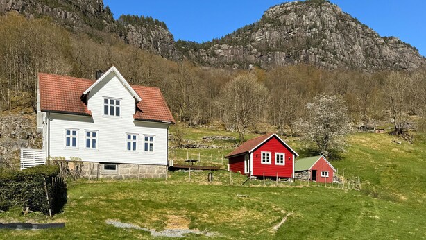Et hvidt hus med rødt tag og en mindre rødstensbygning i et landligt område med et bjerg i baggrunden.