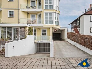 Un immeuble à plusieurs étages doté d’une entrée sécurisée par un portail, d’un balcon aménagé avec du mobilier d’extérieur et surplombant un ciel dégagé.