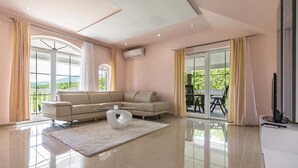A modern living room with a beige sectional sofa, a glass coffee table, and a large window offering a view of greenery.