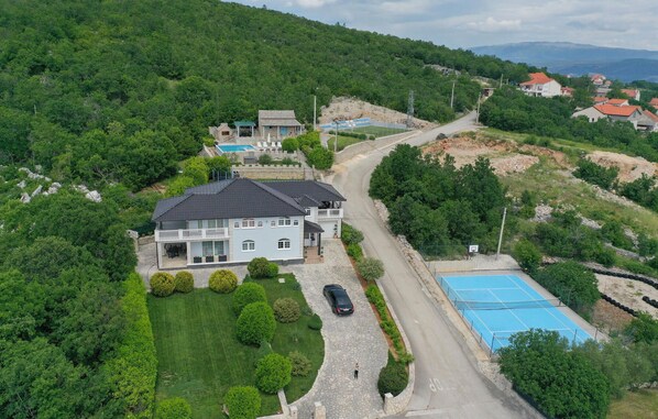 A two-story house with a swimming pool, surrounded by lush greenery and a winding road.