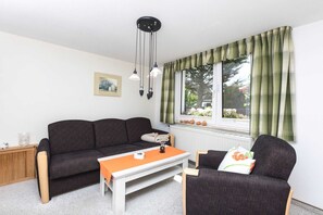 Un salon avec un canapé de couleur sombre, une table basse blanche et un fauteuil avec un coussin. Il y a une fenêtre avec des rideaux verts, un radiateur en dessous et un tableau encadré au mur.