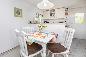 Un coin repas avec une table blanche, deux chaises blanches dotées de coussins marron, une nappe à motifs floraux et un vase rempli de fleurs.