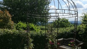 Um gazebo no jardim com um banco, cercado por um caminho de cascalho e vegetação exuberante.