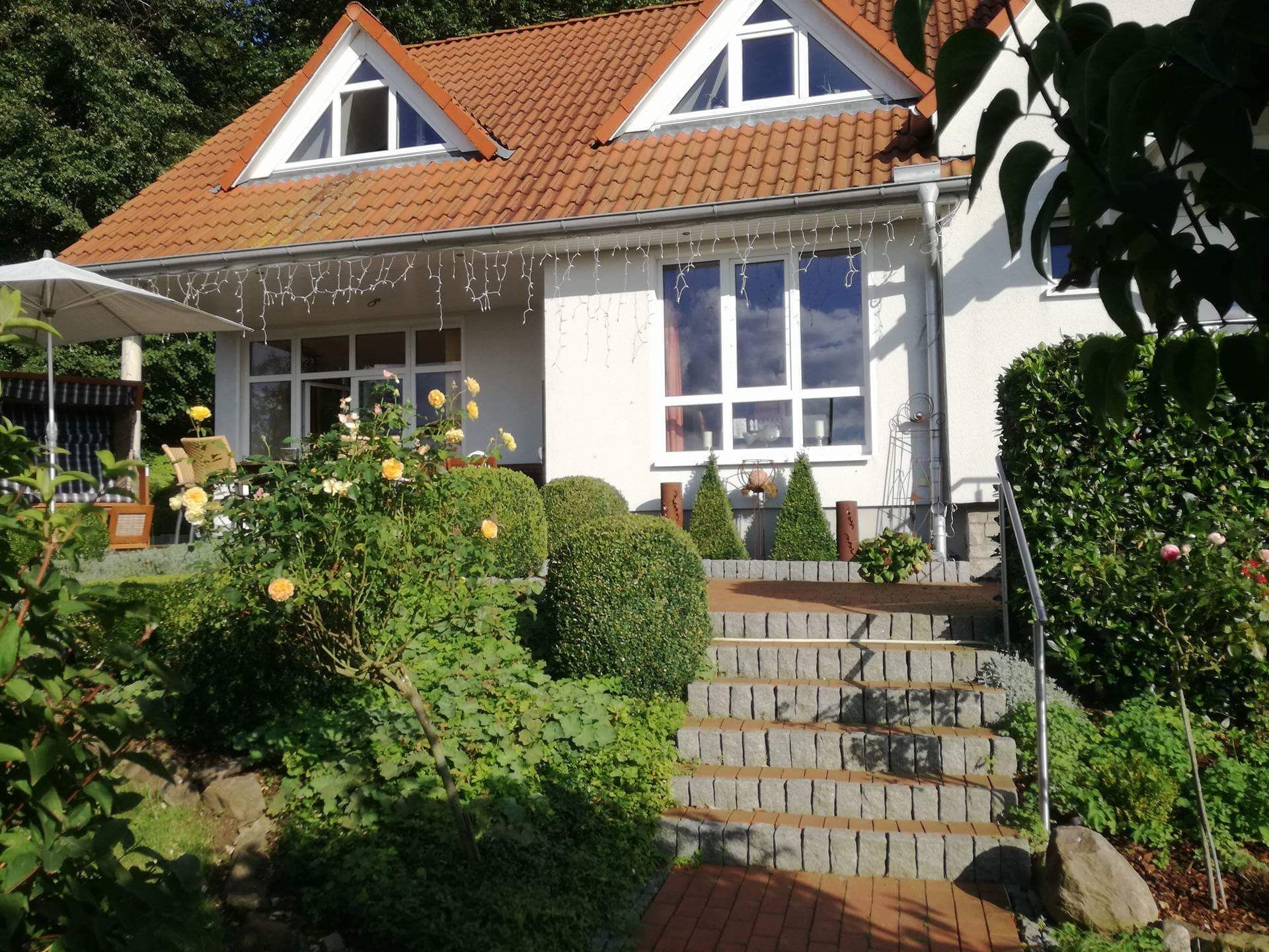 A two-story house with a red-tiled roof, white exterior walls, and a front porch with a white umbrella. There are steps leading up to the porch, and the house is surrounded by greenery and bushes.