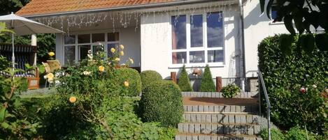 A two-story house with a red-tiled roof, white exterior walls, and a front porch with a white umbrella. There are steps leading up to the porch, and the house is surrounded by greenery and bushes.