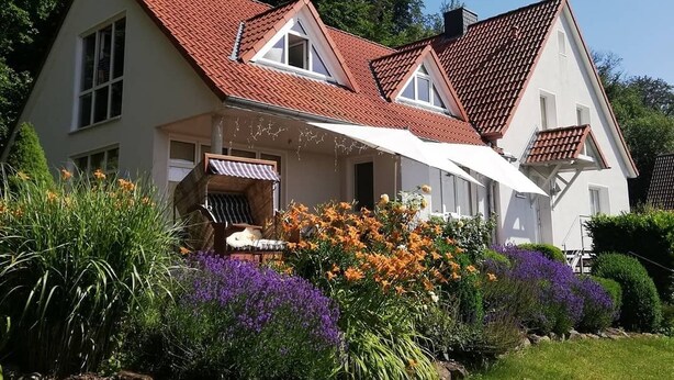 Uma casa de dois andares com telhado de telhas vermelhas, varanda coberta e jardim com diversas flores e plantas.