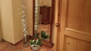 Um corredor com porta de madeira, espelho refletindo um quarto e planta em vaso com luzes.