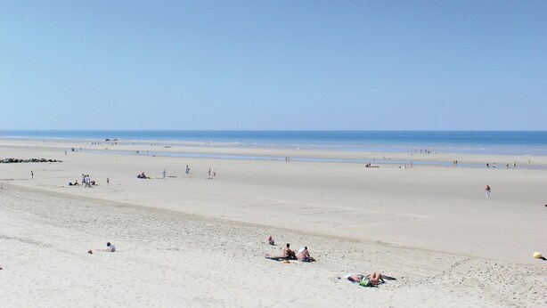 Een breed zandstrand met mensen verspreid over het strand, sommigen liggen op handdoeken en anderen wandelen.
