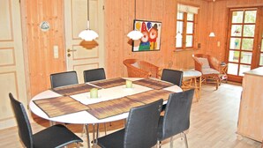 Een eethoek met een ronde tafel, zwarte stoelen en een rieten stoel. Er hangen twee lampen en er is een schilderij van clowns aan de muur.