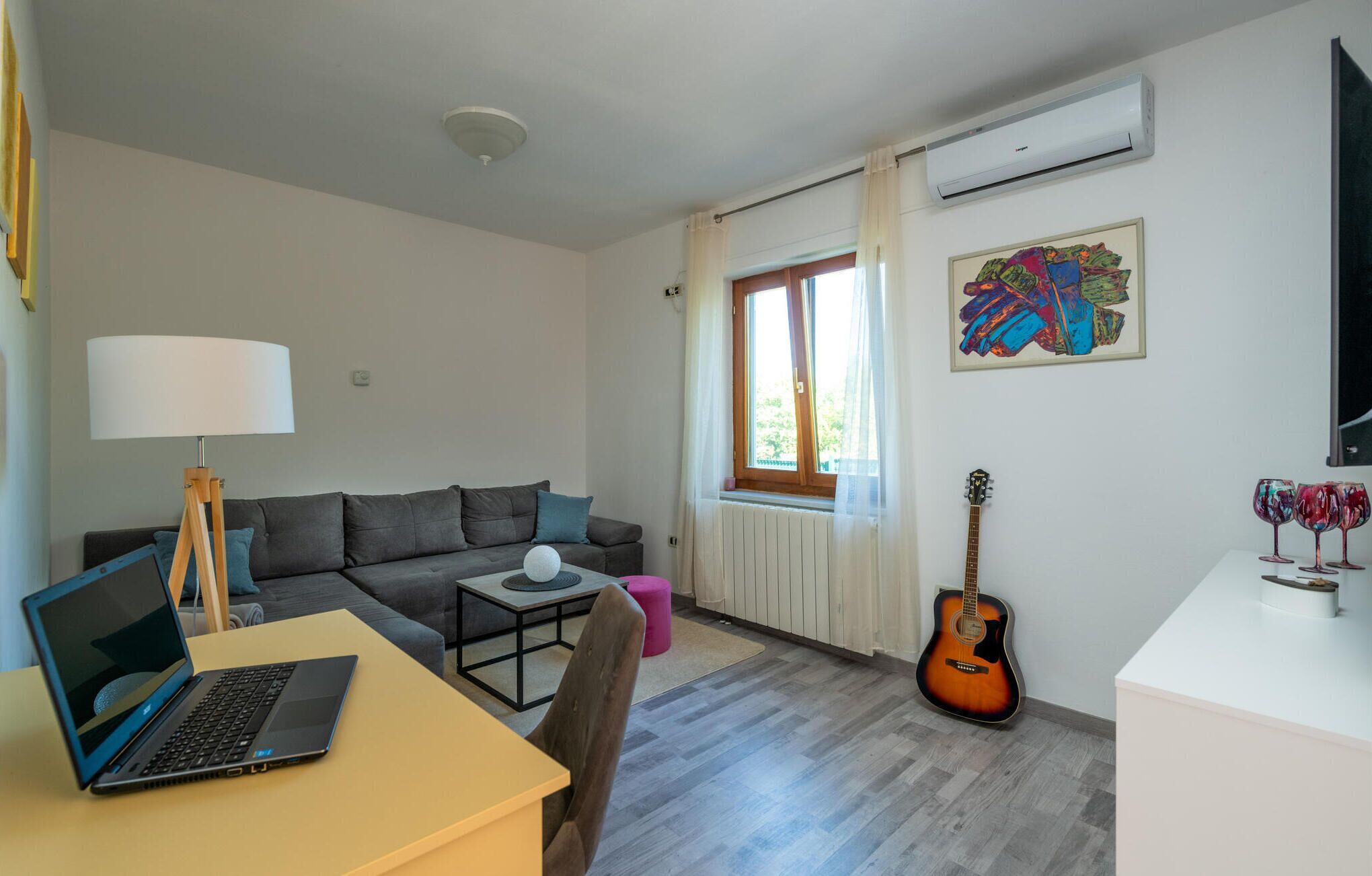 A modern living room with a sofa, a coffee table, a laptop on a desk, a guitar, and a window with curtains.
