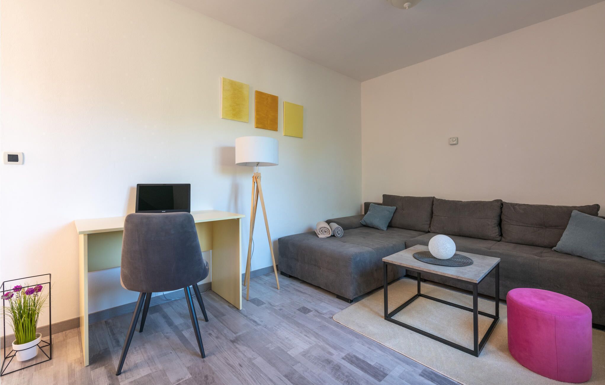 A modern living room with a grey sofa, a small coffee table, a desk with a computer, a floor lamp, and a pink ottoman.