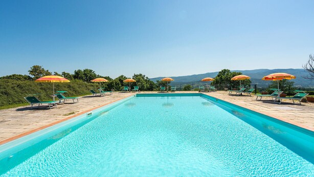 A rectangular swimming pool with clear blue water, surrounded by green lounge chairs and orange umbrellas. - Awesome apartment in Poppi AR with house a panoramic view (Poppi AR)
