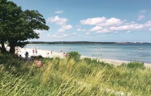 Una playa con gente nadando y caminando por la orilla, rodeada de vegetación y un cielo despejado.