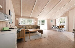 A cozy cabin interior with a wooden floor, a sofa, a dining table with chairs, and large windows offering a view of the outdoors. - 3 bedroom awesome home in Nexø (Nexø)