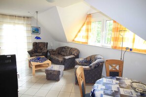 A cozy living room with a high ceiling, a wooden table, patterned armchairs, and a view of the outdoors through a window with orange curtains.