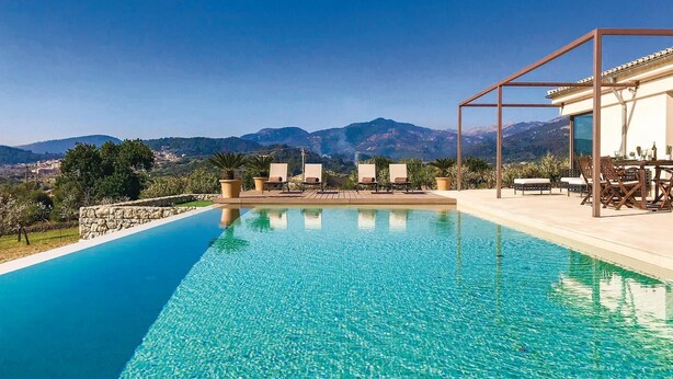 Ein klares, blaues Schwimmbecken mit Liegestühlen und einem Tisch, umgeben von Bergen und viel Grün.