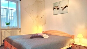 A bedroom with a wooden bed, a nightstand with a lamp, a window with a plant, and a wall with a painting. - (124) 2-room apartment beach road - Appartementhaus Residenz Strandstr. 31 (Kühlungsborn)