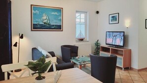 A living room with a sofa, chairs, a dining table, a TV, and a painting of a sailboat. - (124) 2-room apartment beach road - Appartementhaus Residenz Strandstr. 31 (Kühlungsborn)