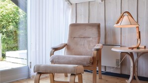 A room with a wooden floor, a beige armchair, a small round table with a lamp, and a window with blinds.