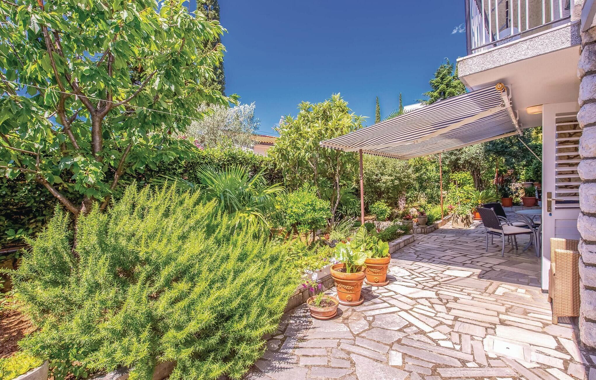 A stone-paved patio with outdoor furniture, surrounded by lush greenery and a pergola providing shade.