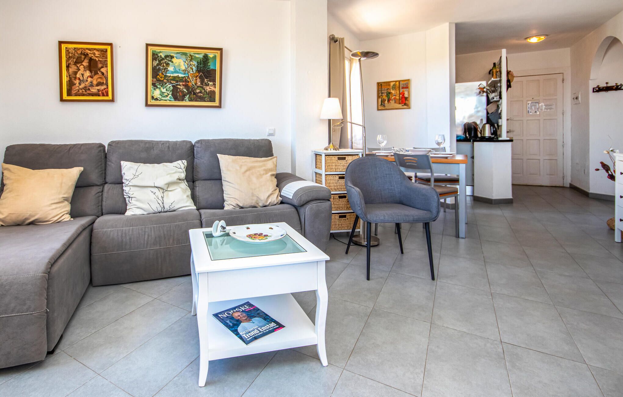 A modern living room with a grey sofa, a white side table with a magazine, and a dining area with a chair and table.