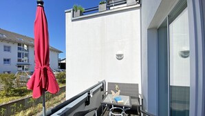 Un balcon agrémenté d’un parasol rouge, une table en verre avec des chaises, et une vue sur un immeuble blanc doté de balcons.