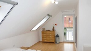 A bedroom with a sloped ceiling, a wooden dresser, a bed with white bedding, a small table, and a window with a view of a red-roofed building. - Semi-detached house / Sonnenstrand No. 5 - Glowe - Semi-detached house Sonnenstrand / (Glowe)