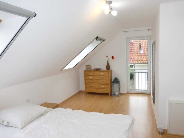 A bedroom with a sloped ceiling, a wooden dresser, a bed with white bedding, a small table, and a window with a view of a red-roofed building.