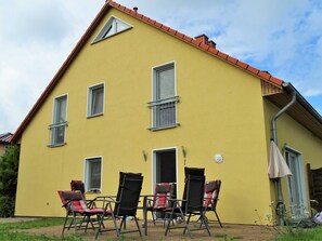 A yellow building with multiple windows, a red roof, and a small garden area with outdoor furniture.