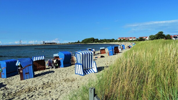 A beach with blue and white striped beach huts, a sandy shore, and a view of the sea and distant buildings. - Semi-detached house / Sonnenstrand No. 5 - Glowe - Semi-detached house Sonnenstrand / (Glowe)