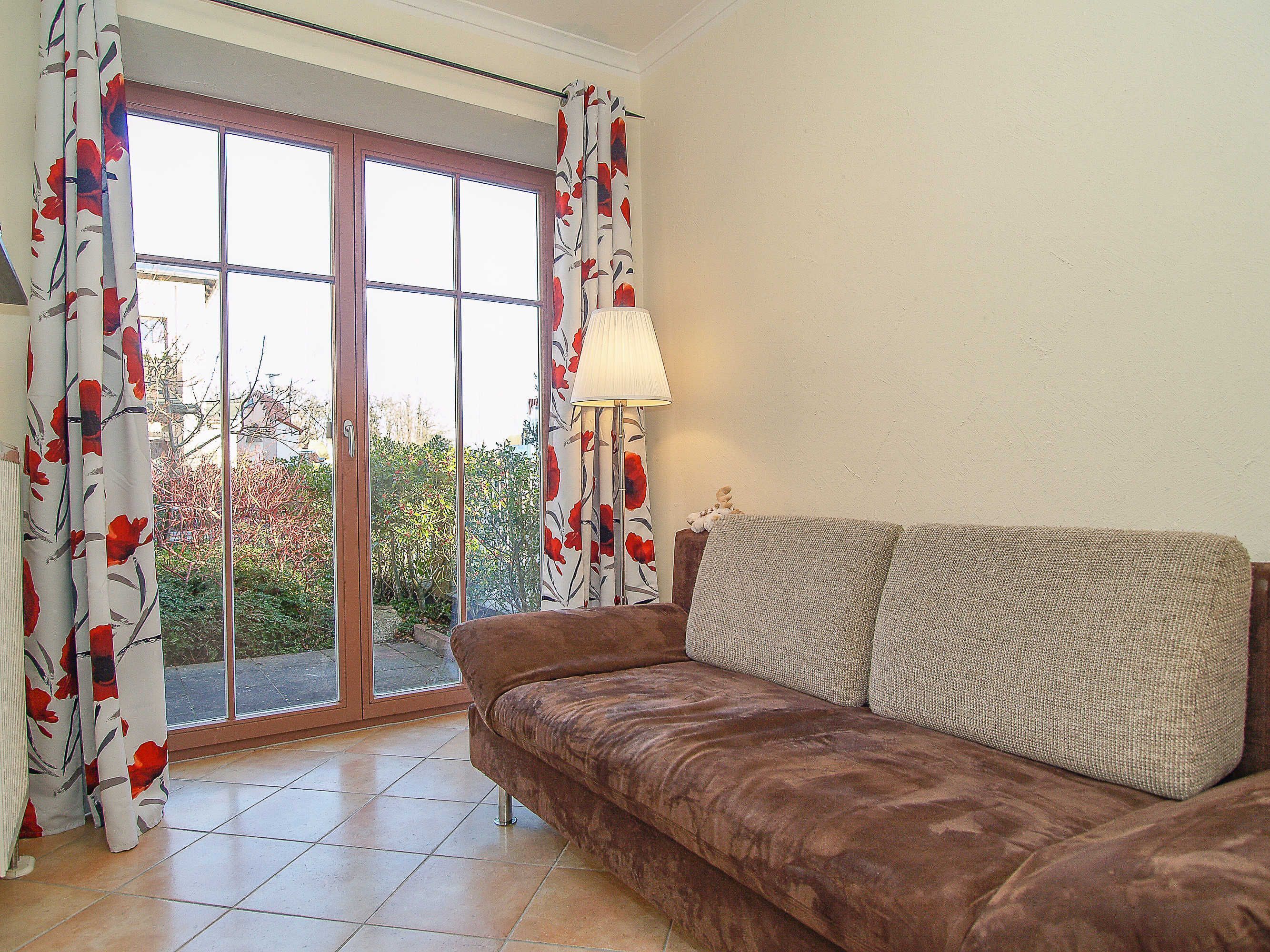 Ein Wohnzimmer mit braunem Sofa, einer Lampe und Blick auf einen Garten durch eine Glastür.