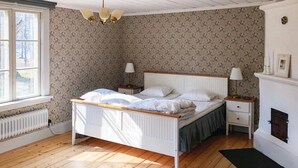 Quarto com cama de madeira, cabeceira branca, duas mesas de cabeceira com luminárias, radiador, lareira e janela com vista para árvores.