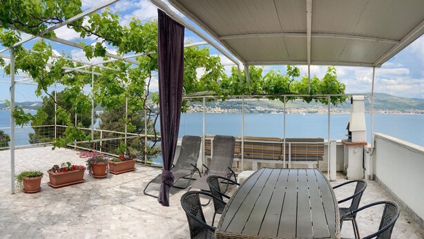 Una terraza con una mesa y sillas, plantas en macetas y vistas al mar y a un pueblo lejano.