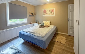A bedroom with a bed, a window, a radiator, a door, and a wall-mounted shelf with a floral picture.