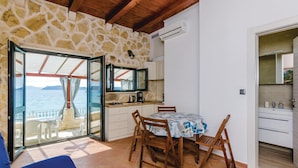 A room with a stone wall, a kitchen area, a dining table with chairs, and a view of the sea through a glass door.