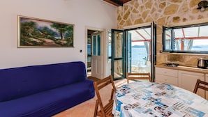 A cozy room with a blue sofa, a painting of a landscape, a kitchen area with a stove, and a view of the sea through open doors.