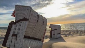 Une grande structure blanche et texturée, qui ressemble à un fauteuil ou à un canapé, se dresse sur une plage de sable, avec un ciel dégagé et l’océan en arrière-plan.