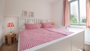 Ein Schlafzimmer mit einem weißen Bett, karierter Bettwäsche, zwei Wandleuchten und einem Fenster mit Vorhängen.
