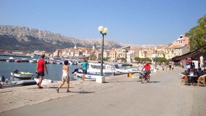 A coastal promenade with people walking and cycling, a marina with boats, and buildings with red-tiled roofs. - Holiday apartment with balcony and air conditioning (Baška)