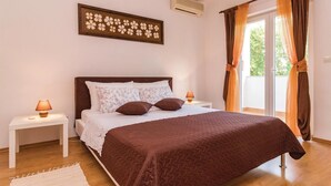 Un dormitorio con cama, dos mesitas de noche, aire acondicionado de pared y ventana con cortinas.