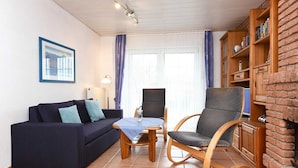 Een woonkamer met een donkerblauwe bank, een houten schommelstoel, een klein houten tafeltje en een boekenkast tegen een bakstenen muur.