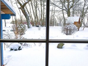 A snowy outdoor scene through a window, including a wooden structure, bare trees, and a snow-covered ground.