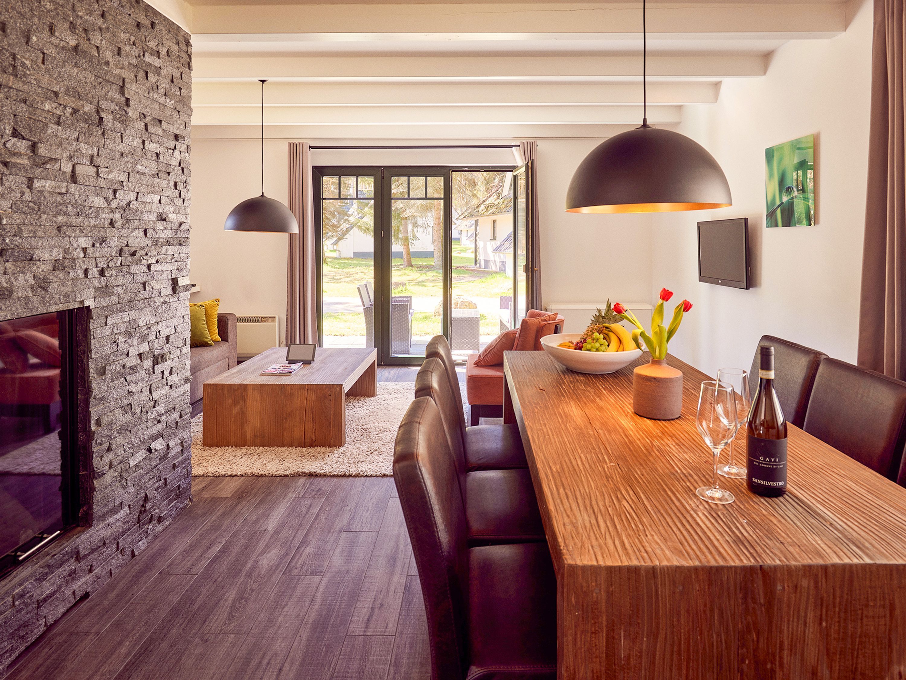 A modern living room with a stone wall, a wooden dining table with a bowl of fruit, and a TV mounted on the wall.
