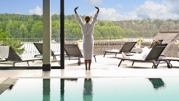 A person in a white robe standing by a pool with a view of a forested hill.
