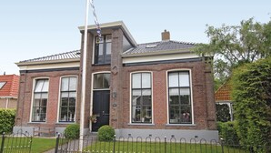Una casa de ladrillo de dos pisos con puerta principal negra, ventanas grandes y un porche pequeño con un banco.