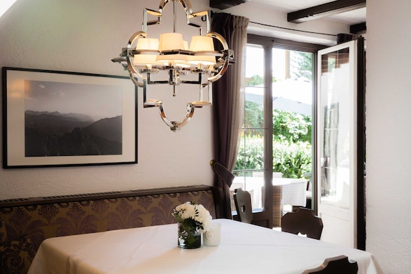 Un coin repas avec une table dressée pour deux personnes, un lustre, un tableau encadré au mur et une fenêtre donnant sur un paysage verdoyant.