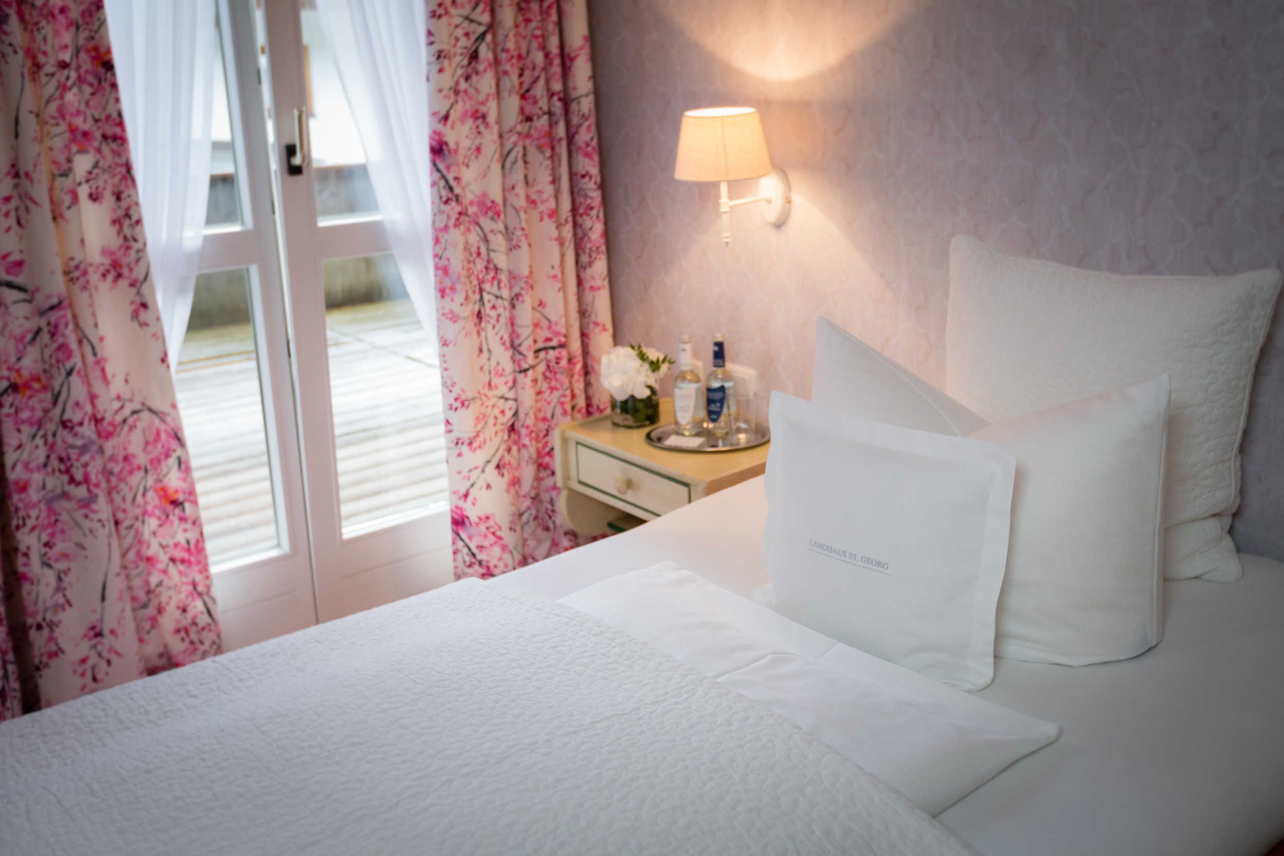 A hotel room with a bed, a nightstand with toiletries, a lamp, and a view of the outdoors through a door.
