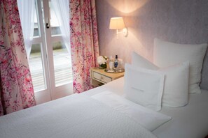 Habitación de hotel con cama, mesita de noche con artículos de aseo, lámpara y vista al exterior a través de una puerta.