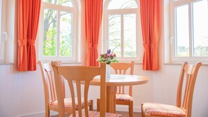 A dining area with a round table and wooden chairs, three windows with arched tops, and orange curtains. - 305 - Hotel Dünenschloss (Karlshagen)